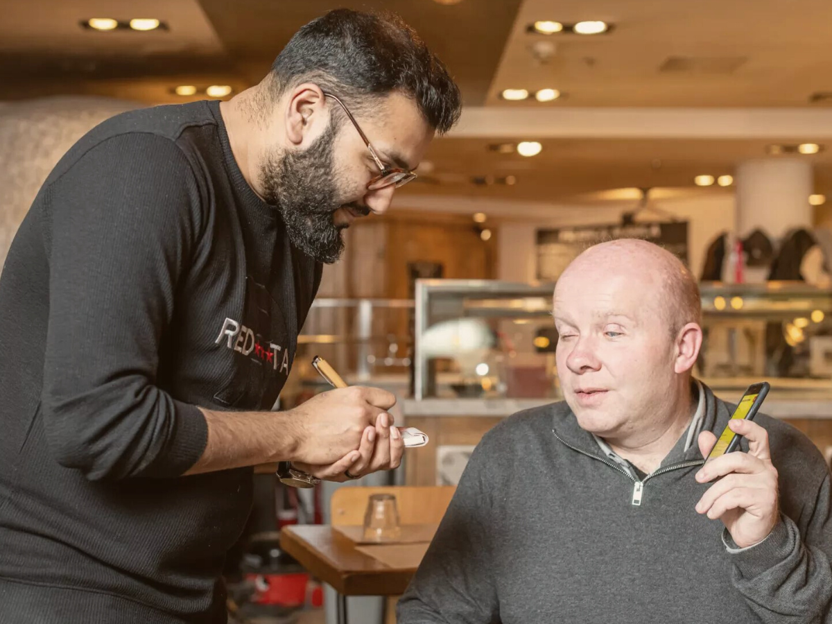 Matt Wadsworth, founder of Good Food Talks who is also visually impaired, listens to his phone that is reading out the menu. A waiter with a notepad and pen waits to hear Matt's order.