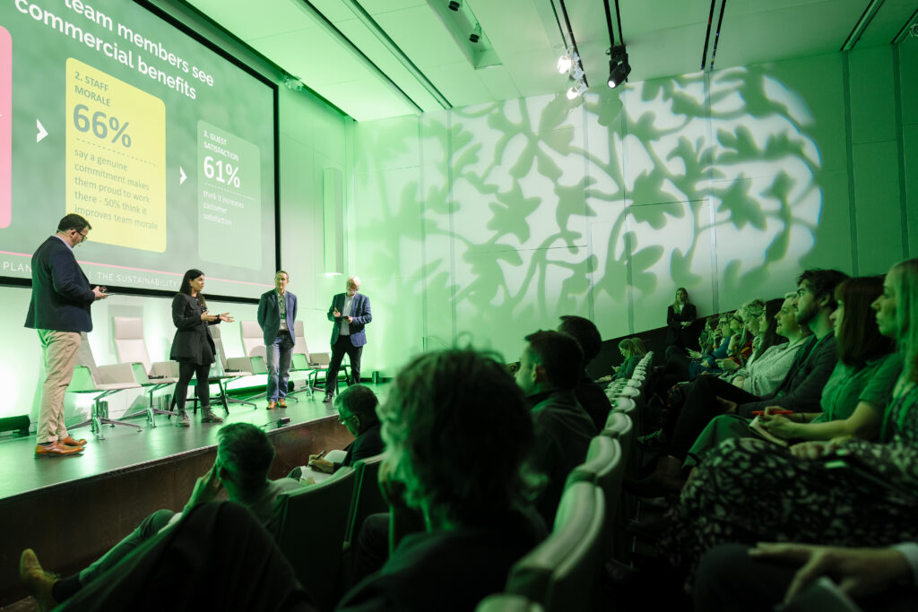 Four people standing on stage at Planet Peach presenting to an audience on commercial benefits.